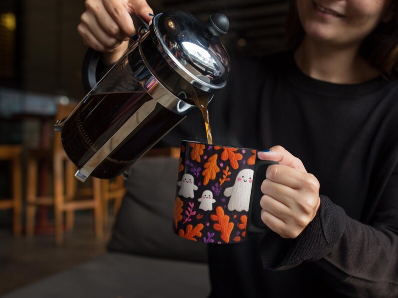 May include: A black coffee mug with a Halloween theme. The mug features white ghost illustrations and orange leaf designs on a black background. Coffee is being poured from a French press into the mug.