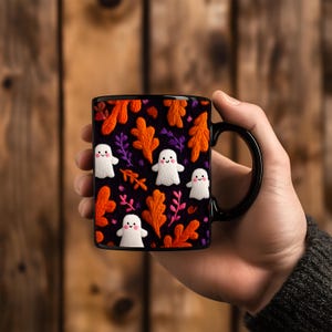 May include: Black ceramic mug featuring a Halloween-themed design. The mug showcases a pattern of white ghost figures, orange and purple leaves, and pink accents against a black background. The mug has a black handle.
