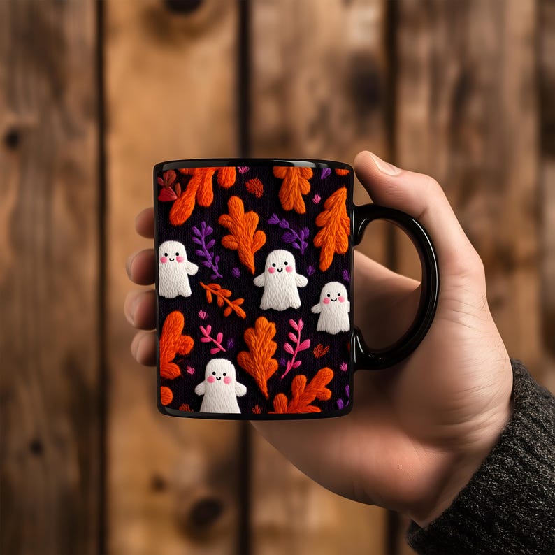 May include: Black ceramic mug featuring a Halloween-themed design. The mug showcases a pattern of white ghost figures, orange and purple leaves, and pink accents against a black background. The mug has a black handle.