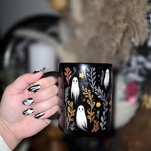 Black ceramic coffee mug featuring a whimsical Halloween design with cute white ghosts surrounded by orange, yellow, and gray autumn foliage and flowers on a dark background. The mug has a glossy finish and a large handle for comfortable use. Available in 11 oz or 15 oz sizes, this festive fall mug is both microwave safe and dishwasher safe, making it a practical yet fun seasonal drinkware choice for coffee, tea, or hot cocoa.
