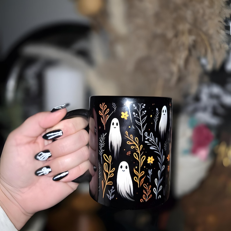 Black ceramic coffee mug featuring a whimsical Halloween design with cute white ghosts surrounded by orange, yellow, and gray autumn foliage and flowers on a dark background. The mug has a glossy finish and a large handle for comfortable use. Available in 11 oz or 15 oz sizes, this festive fall mug is both microwave safe and dishwasher safe, making it a practical yet fun seasonal drinkware choice for coffee, tea, or hot cocoa.