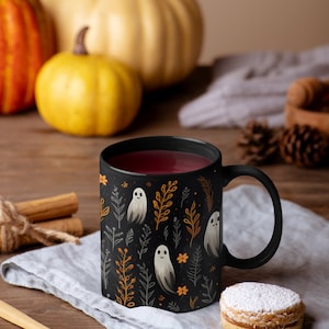 Black ceramic coffee mug featuring a whimsical Halloween design with cute white ghosts surrounded by orange, yellow, and gray autumn foliage and flowers on a dark background. The mug has a glossy finish and a large handle for comfortable use. Available in 11 oz or 15 oz sizes, this festive fall mug is both microwave safe and dishwasher safe, making it a practical yet fun seasonal drinkware choice for coffee, tea, or hot cocoa.