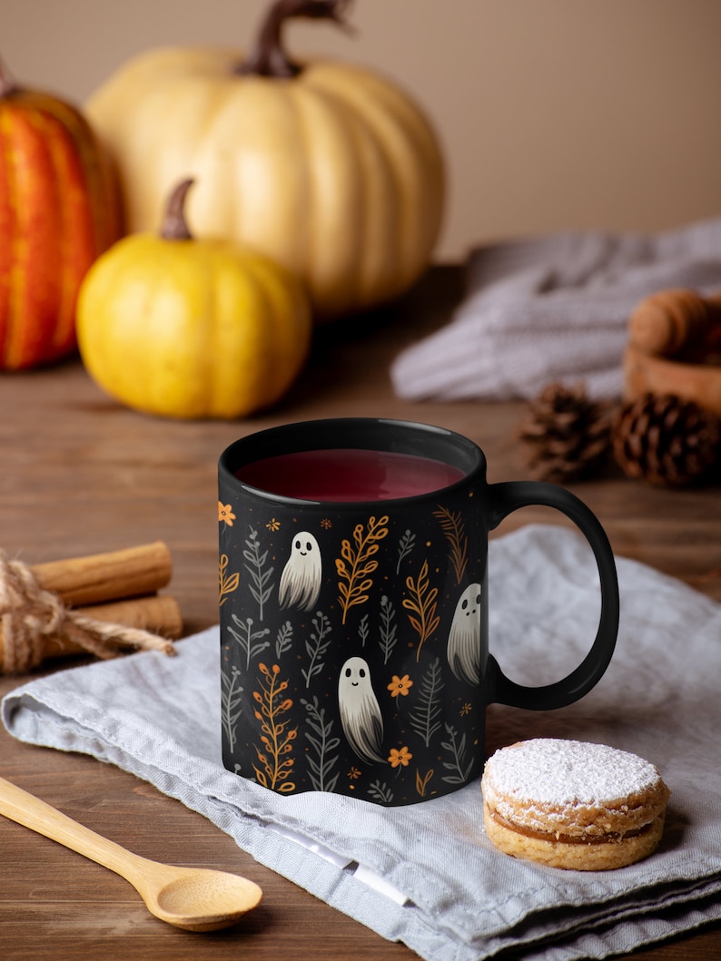 Black ceramic coffee mug featuring a whimsical Halloween design with cute white ghosts surrounded by orange, yellow, and gray autumn foliage and flowers on a dark background. The mug has a glossy finish and a large handle for comfortable use. Available in 11 oz or 15 oz sizes, this festive fall mug is both microwave safe and dishwasher safe, making it a practical yet fun seasonal drinkware choice for coffee, tea, or hot cocoa.
