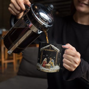 May include: A black coffee mug with a Christmas nativity scene design. The mug is filled with coffee and a person is pouring coffee from a French press.