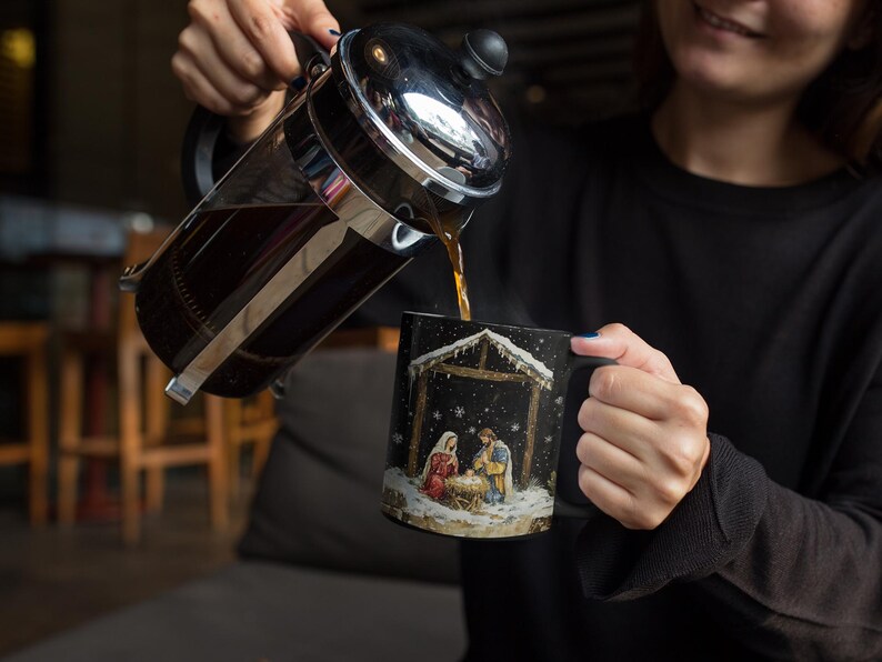 May include: A black coffee mug with a Christmas nativity scene design. The mug is filled with coffee and a person is pouring coffee from a French press.