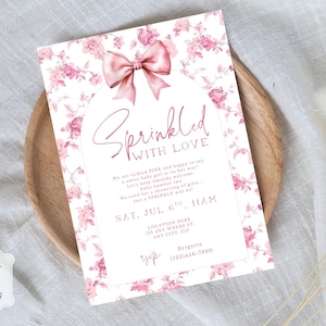 May include: A baby shower invitation with a floral design in pink hues. The invitation features a pink bow and the words "Sprinkled with Love." Details include the date, time, and location. The invitation is on a wooden tray.