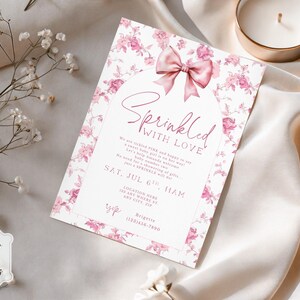 May include: A baby shower invitation with a floral design and a pink bow. The invitation reads "Sprinkled with Love" and includes event details. The invitation is surrounded by white flowers and a candle.