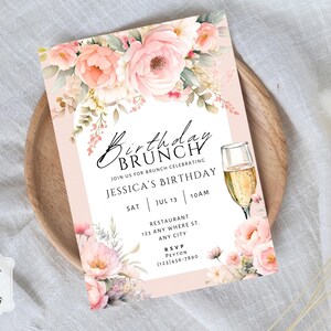 May include: A rectangular birthday brunch invitation card featuring watercolor illustrations of pink and peach flowers at the top and bottom. The text reads Birthday Brunch, Join us for brunch celebrating Jessica's Birthday, Sat Jul 13 10am, Restaurant 123 Any Where St, Any City, RSVP Peyton (123)456-7890, and includes a graphic of a champagne flute.