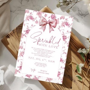 May include: A baby shower invitation with a floral design and a pink bow. The invitation reads "Sprinkled with Love" in elegant script. The invitation includes event details such as date, time, and location. The invitation is on a wooden surface.