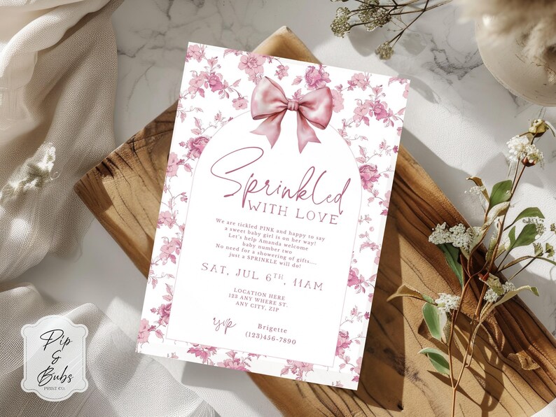 May include: A baby shower invitation with a floral design and a pink bow. The invitation reads "Sprinkled with Love" in elegant script. The invitation includes event details such as date, time, and location. The invitation is on a wooden surface.