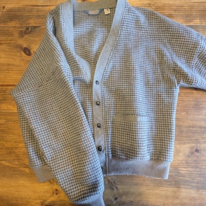 Op de afbeelding: Een grijze cardigan met een pied-de-poule patroon. Het heeft een V-hals, een knoopsluiting en twee voorzakken. De manchetten en zoom zijn grijs geribbeld. De kraag en knooplijst zijn effen grijs.