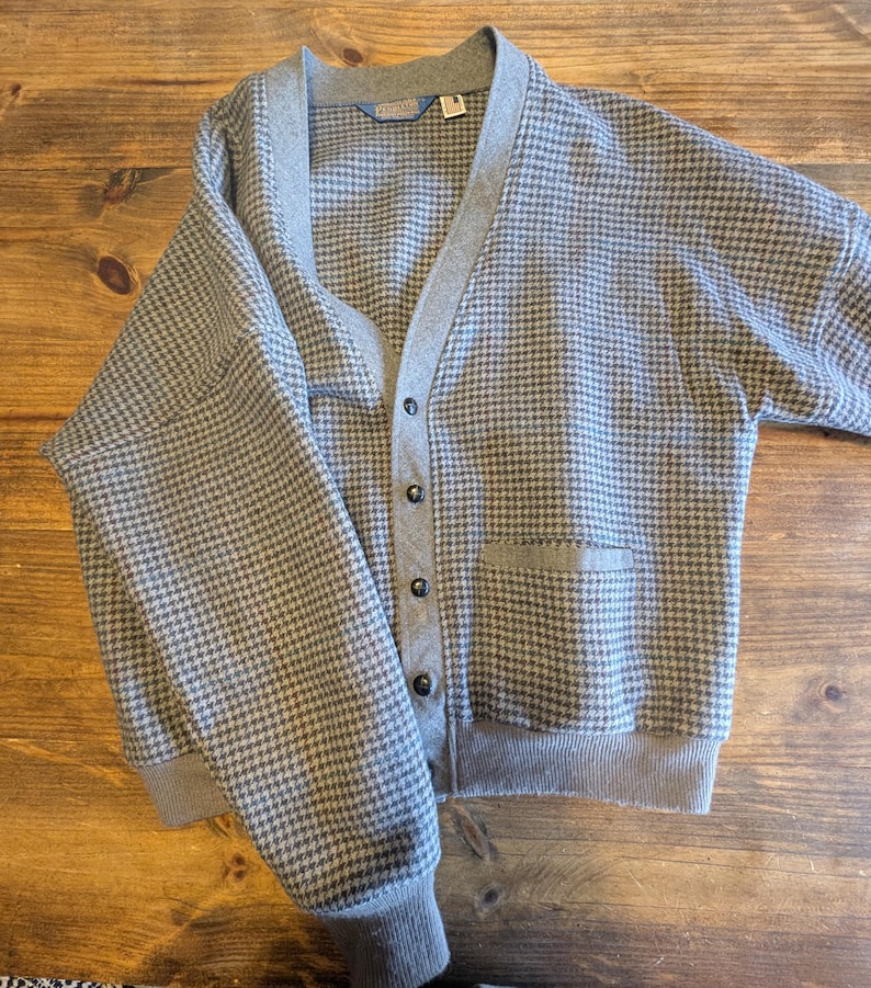 Op de afbeelding: Een grijze cardigan met een pied-de-poule patroon. Het heeft een V-hals, een knoopsluiting en twee voorzakken. De manchetten en zoom zijn grijs geribbeld. De kraag en knooplijst zijn effen grijs.