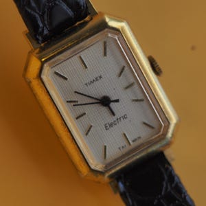 May include: A vintage gold-toned Timex Electric watch with a rectangular face. The watch features a light-colored dial with black hands and markers. The black leather band is visible. The watch face reads "TIMEX" and "Electric".