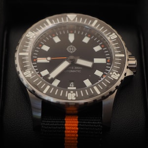 Puede incluir: Un reloj de pulsera negro y plateado con esfera negra, marcadores blancos y detalles en naranja. El reloj tiene un bisel giratorio y una correa a rayas negras y naranjas. La esfera muestra el número 4.