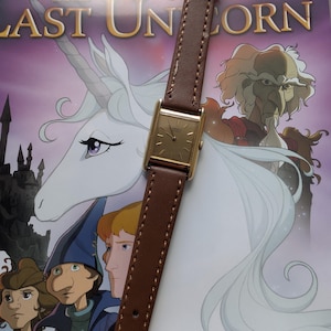 May include: A gold-toned rectangular wristwatch with a brown leather band. The watch is set against a backdrop of a book cover featuring a unicorn and the title "The Last Unicorn". The watch face has a gold color.