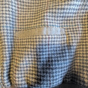 Op de afbeelding: Close-up van een grijze stof met een hondenpoottjespatroon en een klein zakje. De stof heeft een subtiel ruitpatroon met hints van blauw en bordeaux. De onderkant is een donkergrijze geribbelde brei.