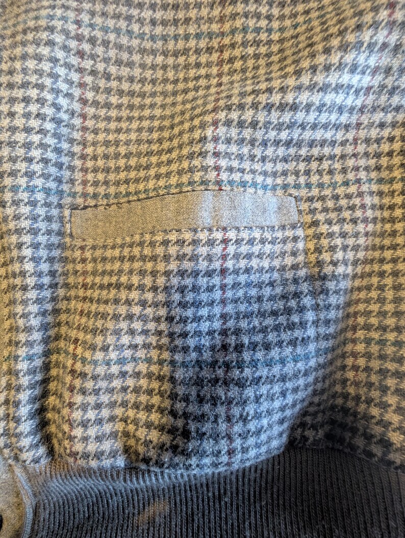 Op de afbeelding: Close-up van een grijze stof met een hondenpoottjespatroon en een klein zakje. De stof heeft een subtiel ruitpatroon met hints van blauw en bordeaux. De onderkant is een donkergrijze geribbelde brei.