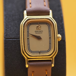 May include: A vintage Seiko Quartz watch with a gold-toned rectangular case and a brown leather strap. The watch face is a light beige color with black hands and the number 12 at the top. The watch is displayed against a dark background.
