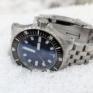Puede incluir: Un reloj de buceo de acero inoxidable con un bisel giratorio negro y una esfera azul oscuro. El reloj presenta marcadores de hora blancos, una pantalla de fecha y una correa de acero inoxidable. El reloj está marcado "DIVER'S 300m AUTOMATIC".