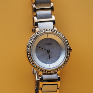 Puede incluir: Un reloj Citizen Eco-Drive con bisel dorado y correa plateada y dorada. La esfera del reloj es gris claro con agujas doradas y el logotipo de Citizen. El bisel está incrustado con pequeños cristales transparentes.