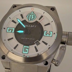 Puede incluir: Un reloj de pulsera automático BALLAST de acero inoxidable. La esfera es blanca con detalles en negro y verde luminoso. Las manecillas y los marcadores de hora del reloj brillan en verde. El reloj tiene una indicación de fecha a las 3 en punto.