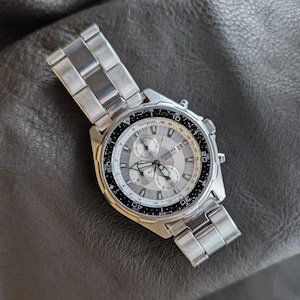 May include: A silver Casio Quartz chronograph watch with a gray dial and three sub-dials. The watch has a black bezel with white markings and a stainless steel band. The watch is displayed on a dark gray surface.