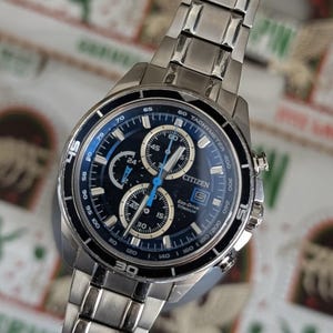May include: A Citizen Eco-Drive titanium watch with a black and blue face. The watch features multiple subdials, a tachymeter, and a silver-tone stainless steel band. The watch face displays the time in both digital and analog formats.