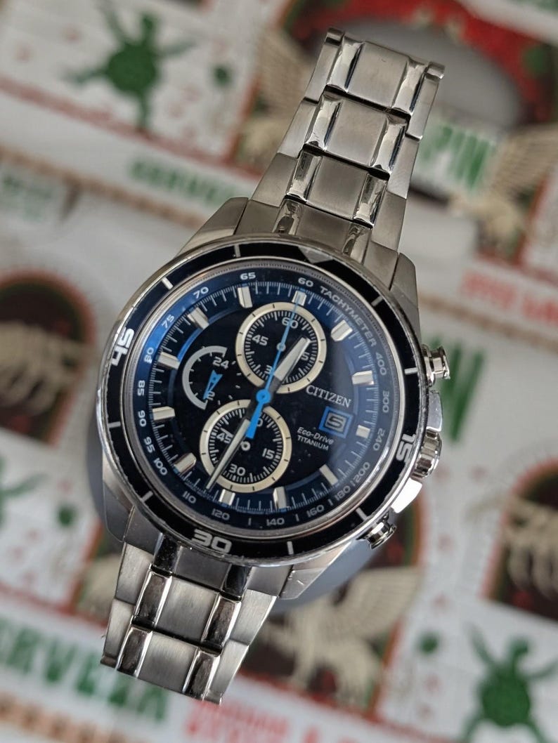 May include: A Citizen Eco-Drive titanium watch with a black and blue face. The watch features multiple subdials, a tachymeter, and a silver-tone stainless steel band. The watch face displays the time in both digital and analog formats.