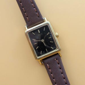 Może przedstawiać: Zegarek Seiko Quartz w kształcie prostokąta w kolorze złotym z czarną tarczą i złotymi wskazówkami. Zegarek ma brązowy skórzany pasek. Tarcza zegarka ma złote znaczniki na każdej godzinie. Zegarek jest w klasycznym stylu vintage.