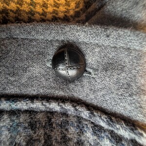 Op de afbeelding: Close-up van een donkergrijze wollen stof met een zwarte, gestikte knoop. De knoop is rond en heeft een kruissteekontwerp. De stof heeft een getextureerd uiterlijk, met een mix van grijze en blauwe draden.