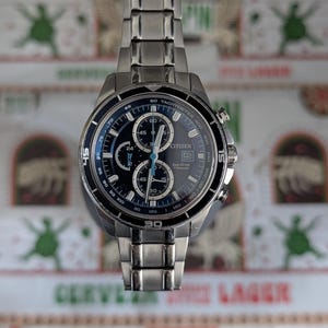 May include: A stainless steel Citizen Eco-Drive titanium chronograph watch with a black dial and blue accents. The watch features multiple sub-dials, a tachymeter bezel, and a stainless steel band. The watch is displayed against a patterned background.
