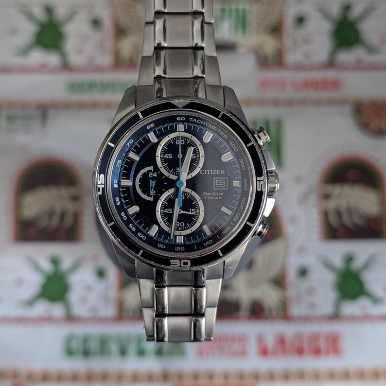 May include: A stainless steel Citizen Eco-Drive titanium chronograph watch with a black dial and blue accents. The watch features multiple sub-dials, a tachymeter bezel, and a stainless steel band. The watch is displayed against a patterned background.