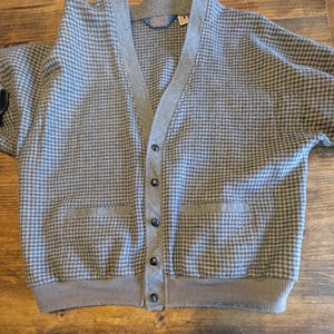 Op de afbeelding: Een grijs en blauw vest met een pied-de-poule patroon, een V-hals, twee voorzakken en een knoopsluiting. De trui heeft een grijze bies en een label bij de hals.