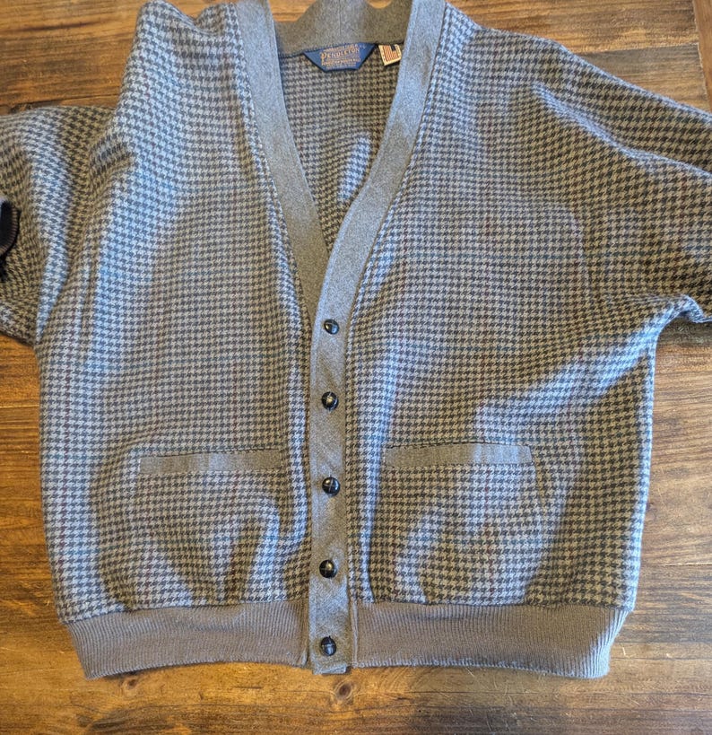 Op de afbeelding: Een grijs en blauw vest met een pied-de-poule patroon, een V-hals, twee voorzakken en een knoopsluiting. De trui heeft een grijze bies en een label bij de hals.