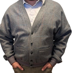 Op de afbeelding: Een grijze cardigan met een pied-de-poule patroon, V-hals, knoopsluiting en twee voorzakken. De cardigan heeft grijze afwerking en manchetten. Een wit overhemd met kraag is eronder zichtbaar.