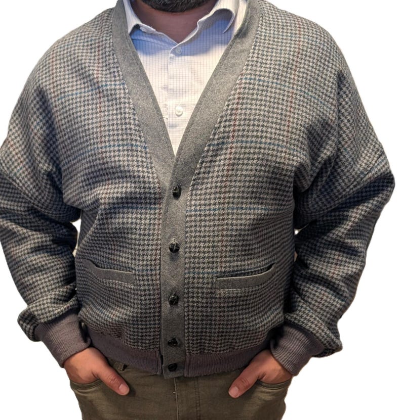 Op de afbeelding: Een grijze cardigan met een pied-de-poule patroon, V-hals, knoopsluiting en twee voorzakken. De cardigan heeft grijze afwerking en manchetten. Een wit overhemd met kraag is eronder zichtbaar.