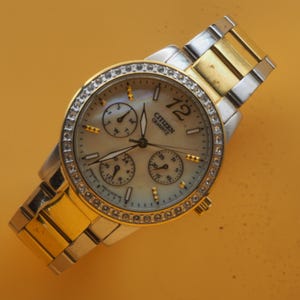 Può includere: Un orologio da polso Citizen Quartz con cinturino oro e argento. Il quadrante è di colore chiaro con tre sotto-quadranti e una lunetta tempestata di diamanti. Il numero 12 è ben visibile.