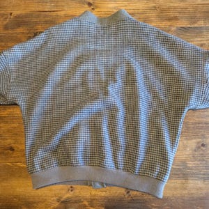 Op de afbeelding: Een grijze en zwarte bomberjack met pied-de-poule patroon en geribde kraag, manchetten en zoom. De jas ligt plat op een houten oppervlak en toont zijn volledige vorm en ontwerp. De stof heeft een klassiek, geweven patroon.