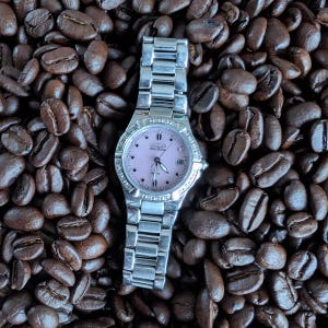 May include: A silver-toned Citizen Eco-Drive watch with a light pink face and a date display, set against a backdrop of dark brown coffee beans. The watch has a silver band and a bezel with small, clear stones.