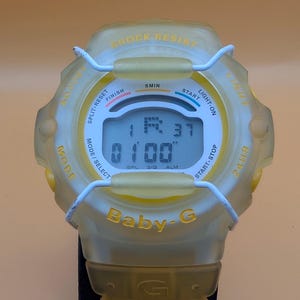 Puede incluir: Un reloj digital Baby-G amarillo con una correa negra. La esfera del reloj muestra la hora y varias funciones. El reloj tiene detalles en blanco y las palabras "SHOCK RESIST" y "Baby-G" son visibles.