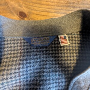 Op de afbeelding: Close-up van een grijze wollen jas kraag met een pied-de-poule patroon. De jas heeft een Pendleton label en een klein Amerikaans vlaggetje, wat aangeeft dat hij in de VS is gemaakt. De stof lijkt van hoogwaardige wol te zijn.