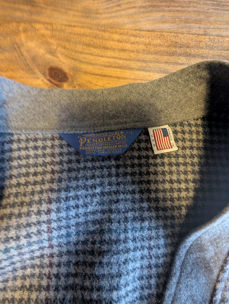 Op de afbeelding: Close-up van een grijze wollen jas kraag met een pied-de-poule patroon. De jas heeft een Pendleton label en een klein Amerikaans vlaggetje, wat aangeeft dat hij in de VS is gemaakt. De stof lijkt van hoogwaardige wol te zijn.