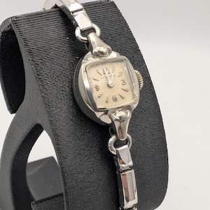 May include: A vintage silver-toned wristwatch with a rectangular face and a cream-colored dial. The watch features silver hour markers and hands. The watch is displayed on a black stand. The watch band is also silver-toned.