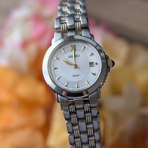 Op de afbeelding: Een zilver- en goudkleurig Seiko-polshorloge met een witte wijzerplaat. Het horloge heeft gouden wijzers en Romeinse cijfers als uurmarkeringen. De datum wordt weergegeven op 3 uur. De horlogeband is in zilver- en goudkleurige schakelstijl.