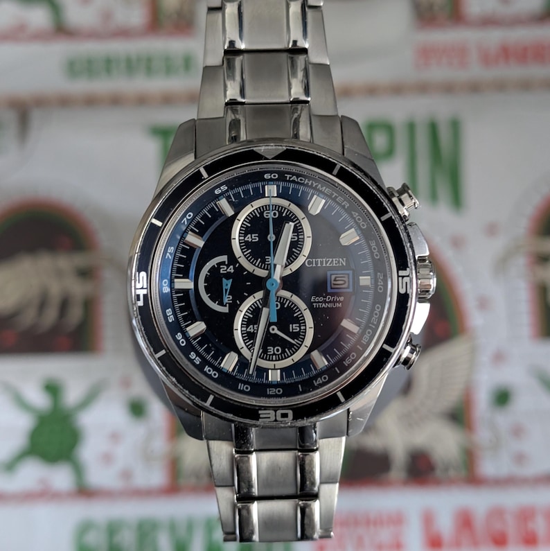 May include: A Citizen Eco-Drive Titanium watch with a dark blue face and silver-tone stainless steel band. The watch features multiple subdials, a tachymeter bezel, and the Citizen logo. The watch is a stylish accessory.
