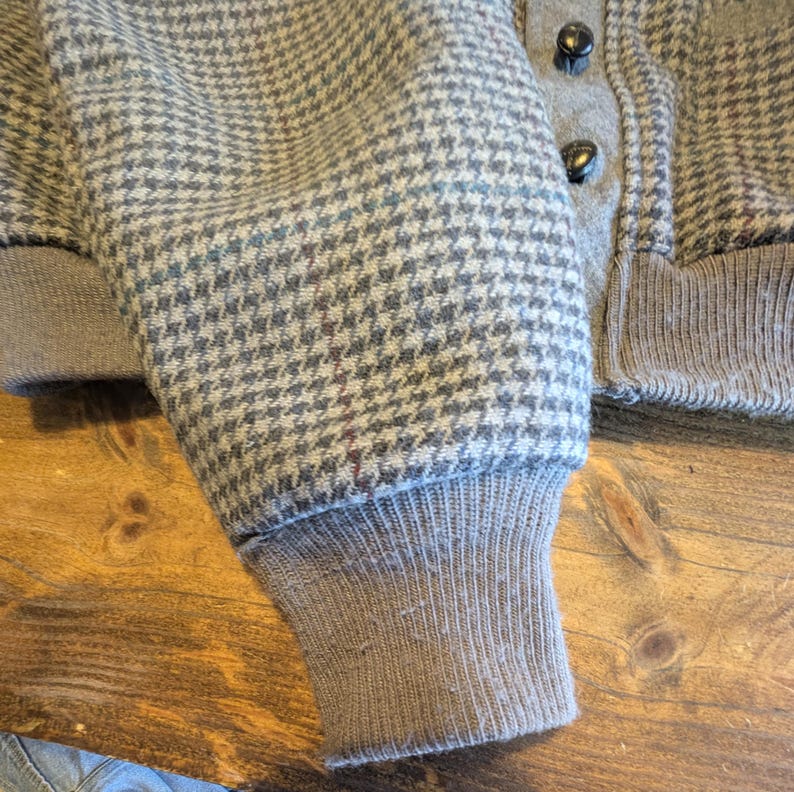 Op de afbeelding: Close-up van een mouw van een jas met grijs en wit pied-de-poule patroon. De mouw heeft een geribde manchet en een knoopsluiting. De jas lijkt gemaakt te zijn van een wolmix.