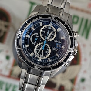 May include: A silver-toned Citizen Eco-Drive Titanium watch with a dark blue face. The watch features multiple sub-dials, a tachymeter, and the Citizen logo. The watch band is made of silver-toned metal links.