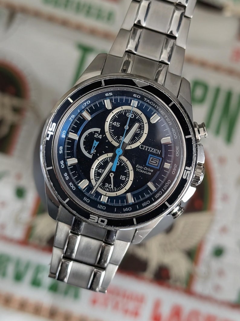 May include: A silver-toned Citizen Eco-Drive Titanium watch with a dark blue face. The watch features multiple sub-dials, a tachymeter, and the Citizen logo. The watch band is made of silver-toned metal links.