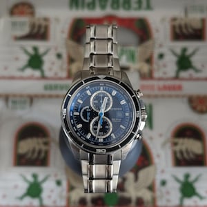 May include: A stainless steel Citizen Eco-Drive chronograph watch with a blue dial. The watch features multiple sub-dials, a tachymeter bezel, and a date display. The watch has a stainless steel band and is displayed on a dark gray stand.