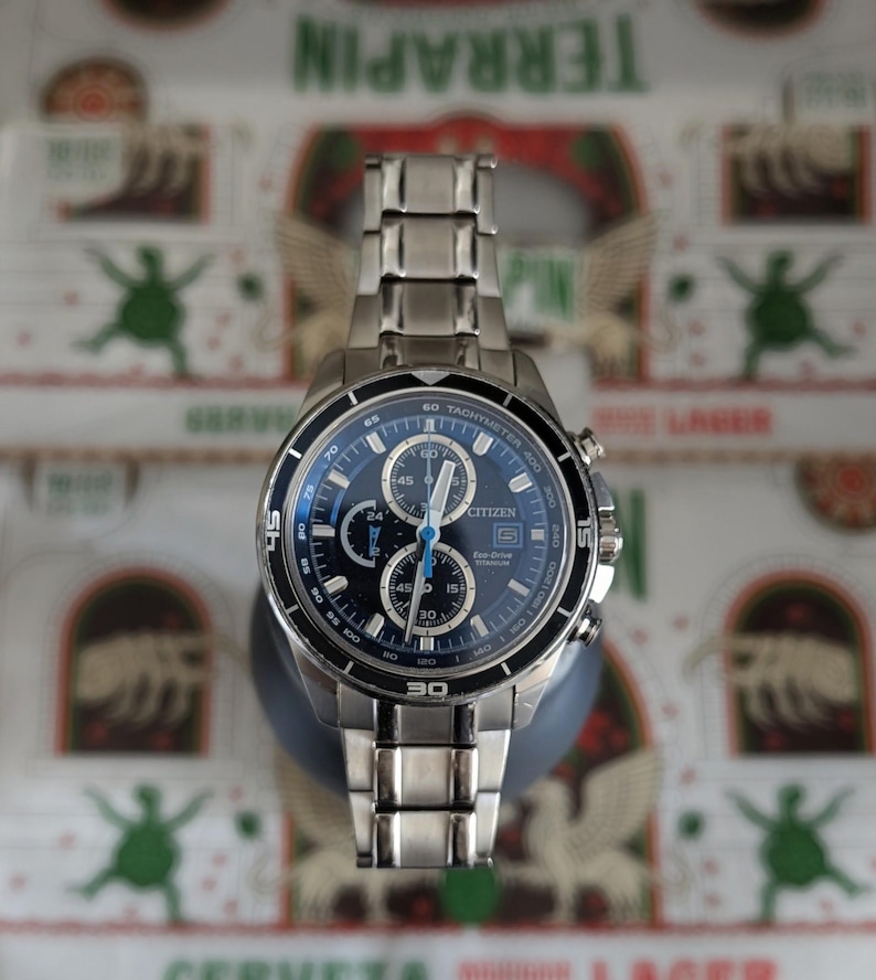 May include: A stainless steel Citizen Eco-Drive chronograph watch with a blue dial. The watch features multiple sub-dials, a tachymeter bezel, and a date display. The watch has a stainless steel band and is displayed on a dark gray stand.
